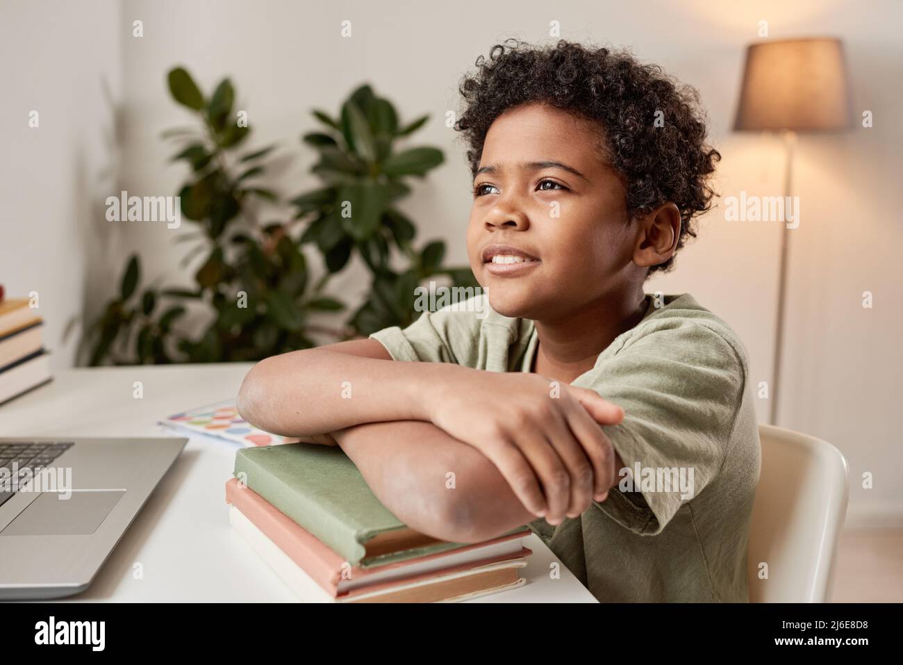 Sorridente ragazzo nero sognante con capelli ricci seduto con le braccia incrociate su pile di libri alla scrivania nella sua camera Foto Stock