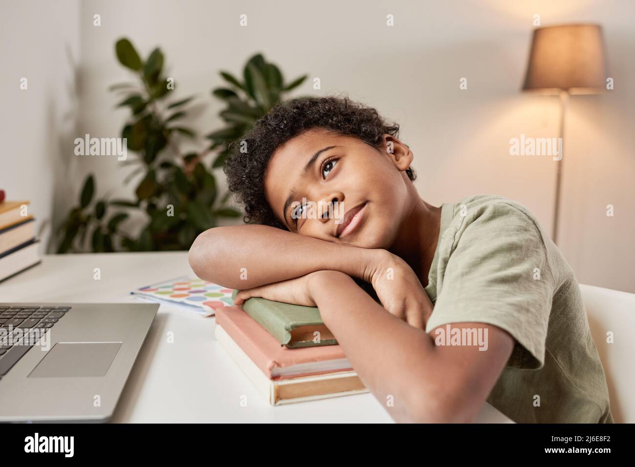 Ragazzo africano carino e pensivo in maglietta seduta al tavolo e sdraiato su una pila di libri mentre si guarda in su Foto Stock
