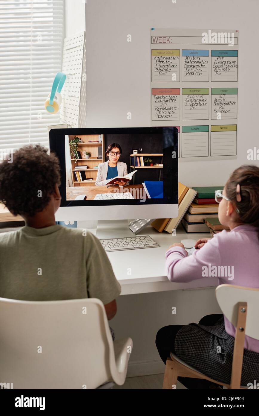 Vista posteriore di bambini multietnici seduti al tavolo con il computer moderno e ascoltando il tutor ispanico online Foto Stock