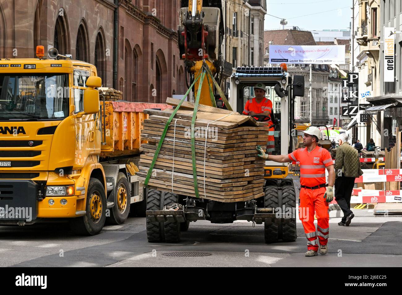 Basilea, Svizzera - Aprile 2022: Il lavoratore edile che aiuta a spostare un carico pesante di legname su una strada urbana con l'aiuto di un digger mobile Foto Stock