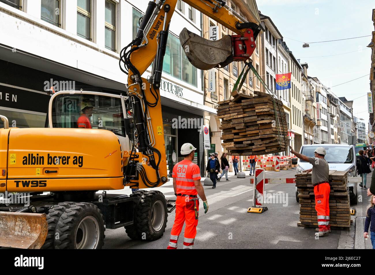 Basilea, Svizzera - Aprile 2022: Il lavoratore edile che aiuta a spostare un carico pesante di legname su una strada urbana con l'aiuto di un digger mobile Foto Stock