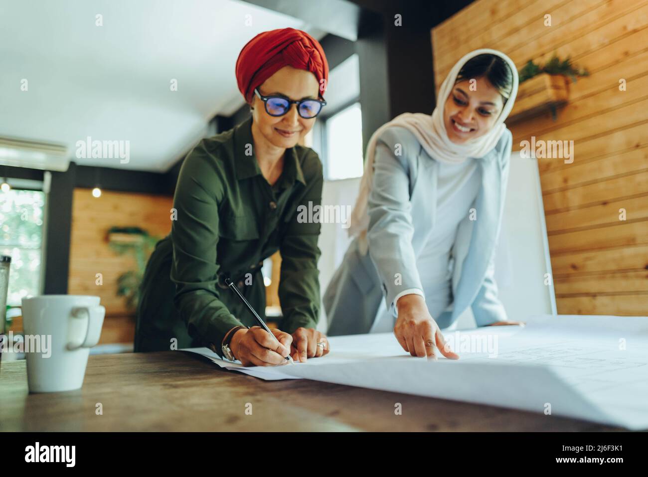 Felici architetti musulmani che lavorano su disegni blueprint in un ufficio moderno. Due imprenditrici creative che progettano un nuovo progetto innovativo. Designazione femminile Foto Stock