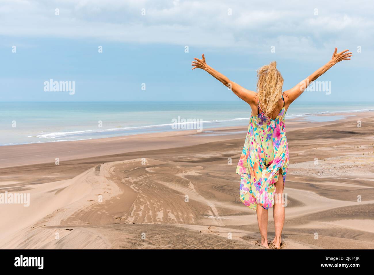 Foto con spazio copia del retro di una donna bionda che alza le braccia sulla parte superiore di una duna di fronte all'oceano Foto Stock