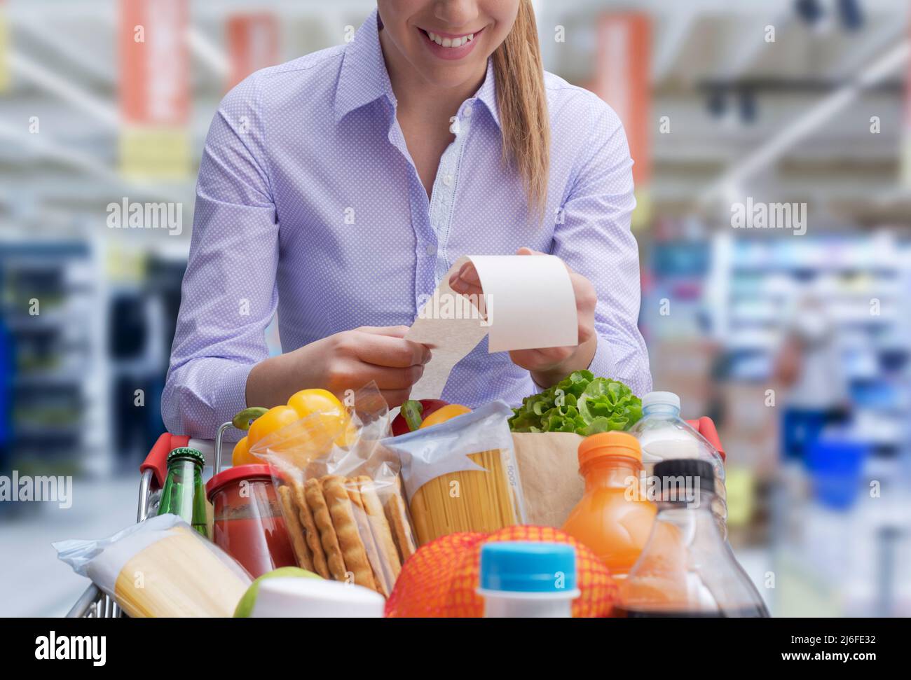 Donna sorridente appoggiata sul carrello della spesa e controllando la ricevuta della drogheria Foto Stock