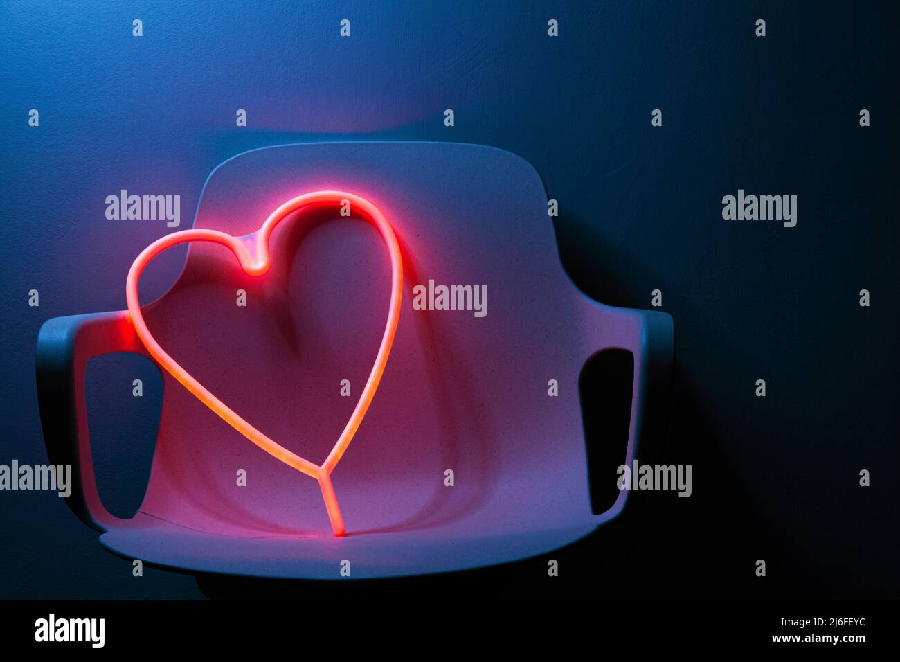 Cuore d'amore rosa al neon su una sedia da ufficio in una stanza blu scuro. Foto Stock
