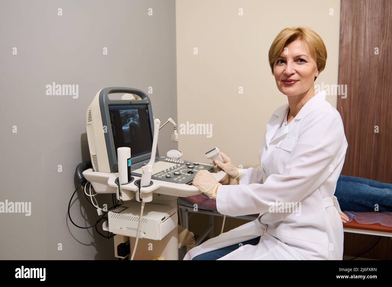 Piacevole donna caucasica adulta, radiologo, specialista di diagnostica, seduto alla macchina a ultrasuoni, tenendo il trasduttore, guardando con sicurezza la fotocamera Foto Stock