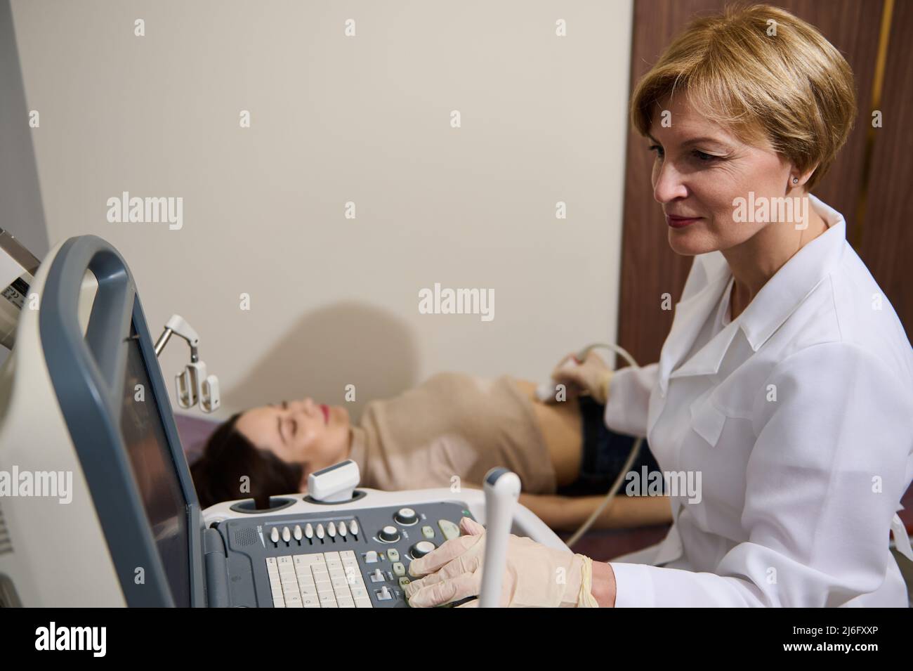 Fidata caucasica adulta medico sposta una sonda di contatto sopra l'addome di una giovane donna paziente sdraiata al daybed durante una visita medica o Foto Stock