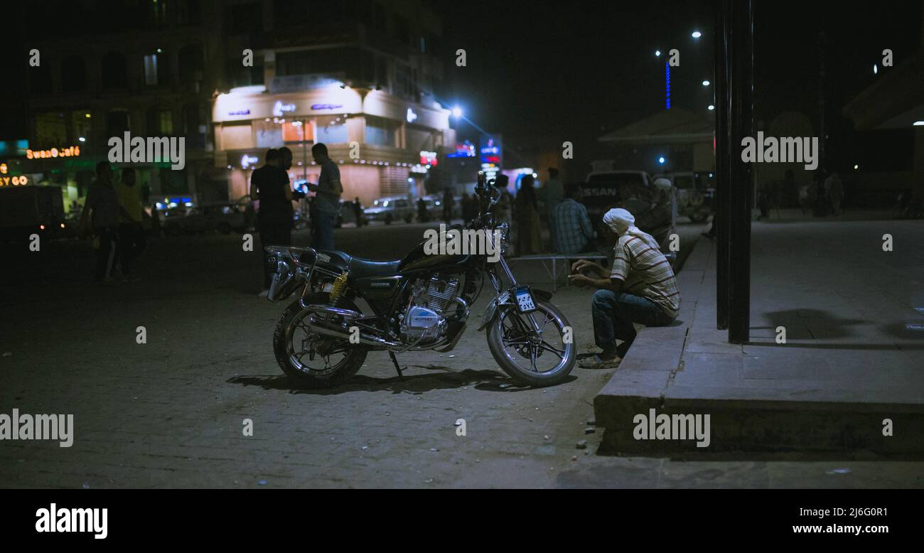 Uomo egiziano seduto presso la stazione ferroviaria di Assuan nuovo alla sua moto di notte. Foto Stock