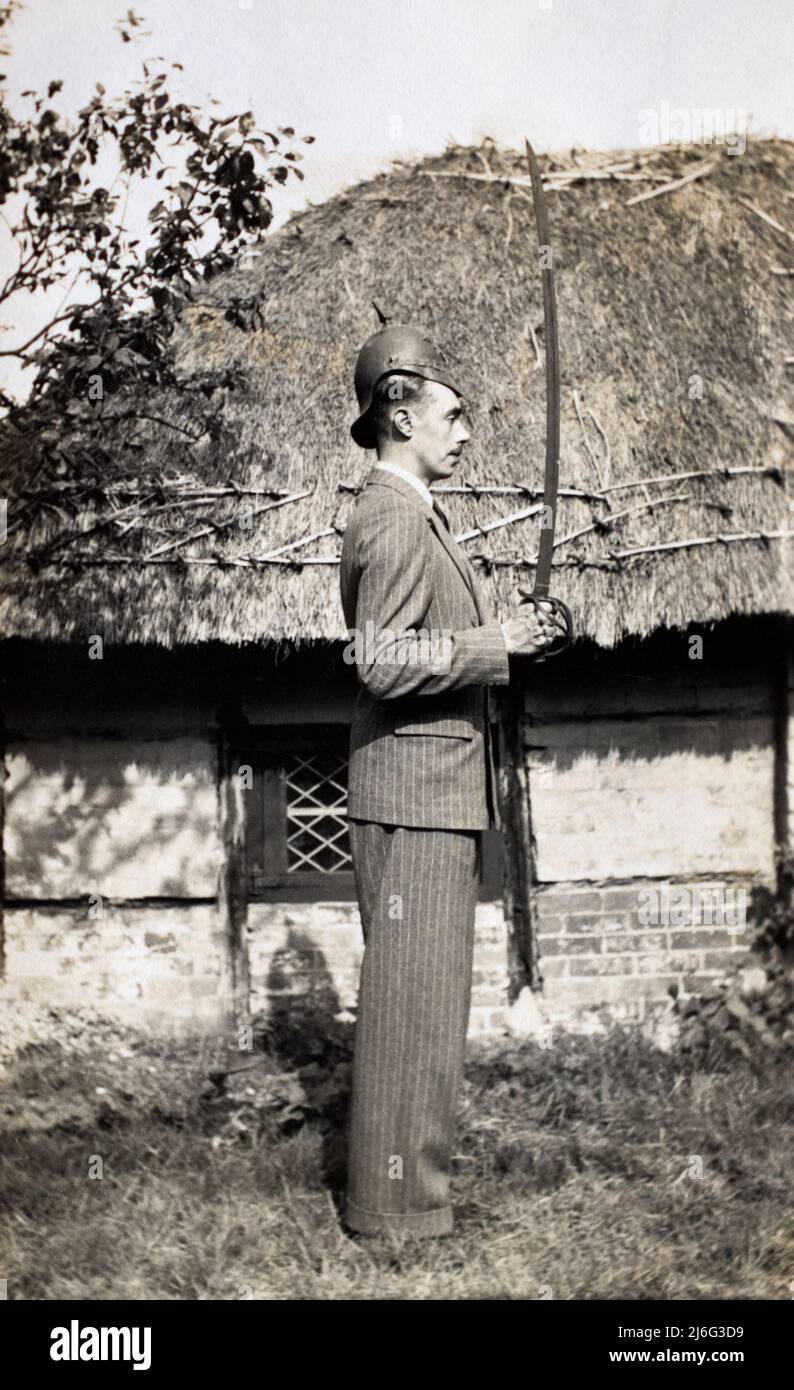 Un uomo in un vestito a strisce spilla che posa fuori di un cottage di paglia. Indossa un vecchio casco di metallo di un reggimento britannico di cavalleria pesante e tiene una spada. Foto Stock