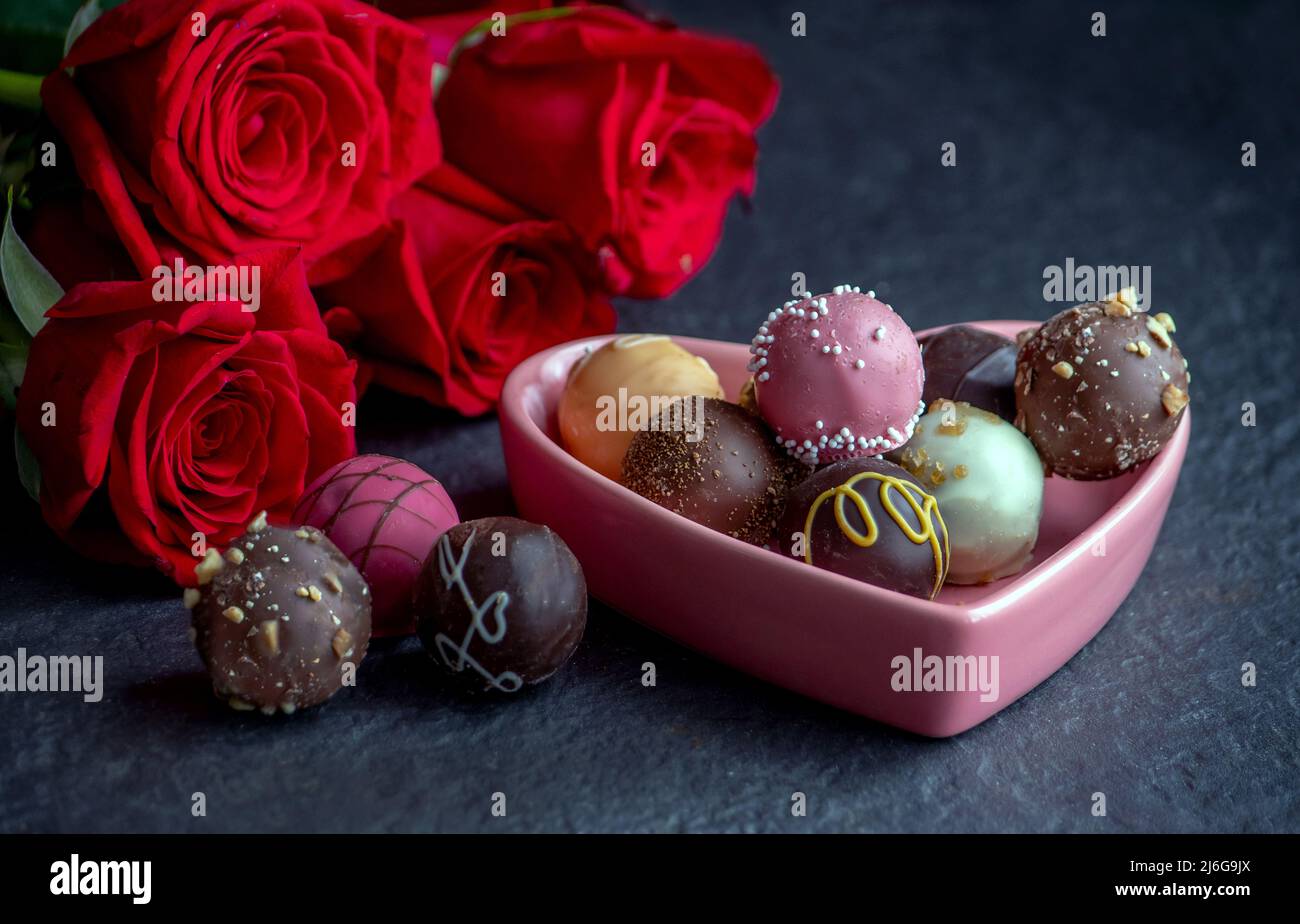 deliziosa varietà di tartufi di cioccolato in un piatto di caramelle a cuore rosa, con rose rosse Foto Stock