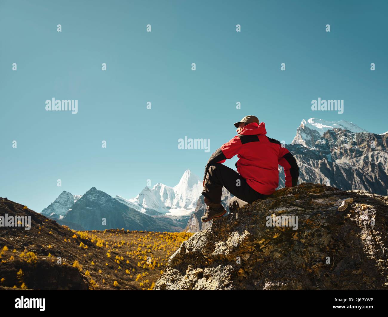 Uomo asiatico seduto in cima alla roccia con Yangmaiyong (o Jampayang in tibetano) picco di montagna in lontananza in Yading, Contea di Daocheng, Provincia di Sichuan Foto Stock