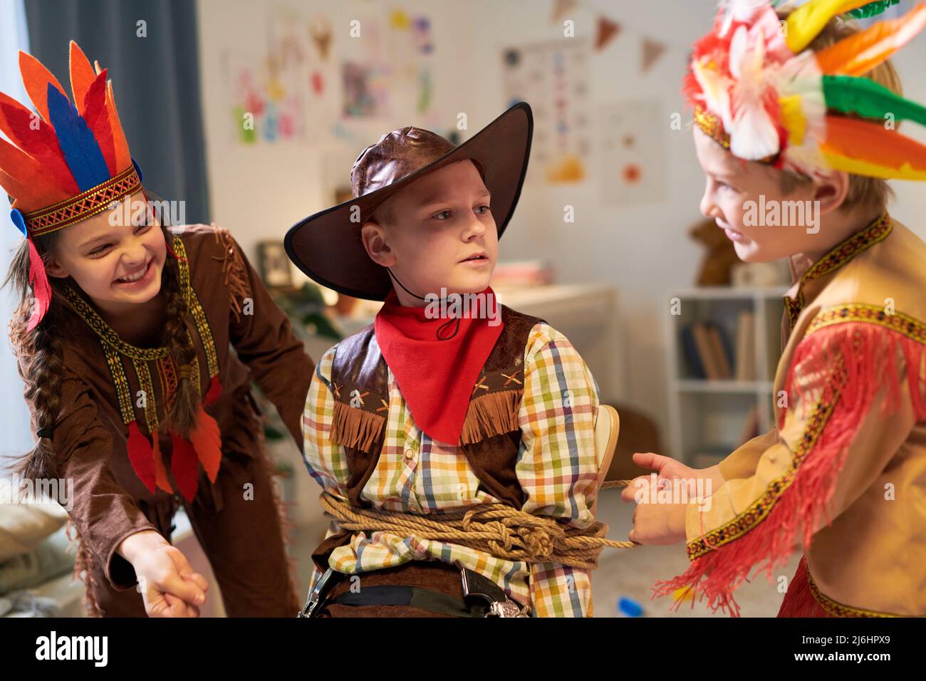 I bambini felici nei costumi del palco che legano il cowboy catturato con la corda mentre giocano ed hanno divertimento durante le riprese commerciali Foto Stock
