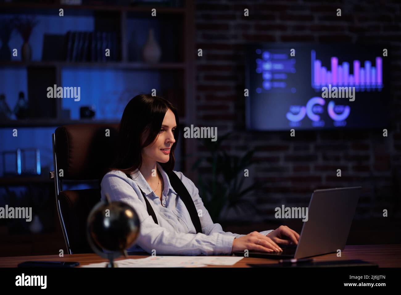 Giovane donna che lavora con un notebook in ufficio. Il lavoratore d'ufficio che analizza grafici e grafici, lavora in modo eccessivo mentre è seduto in ufficio fino a tardi. Cinematografico Foto Stock