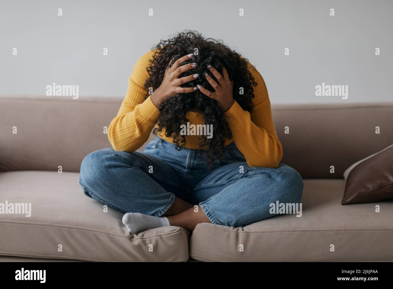 Depressa giovane donna nera seduta a gambe incrociate sul divano con la testa in mano a casa Foto Stock