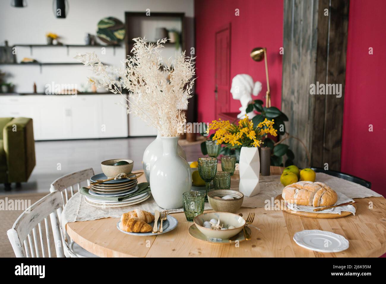Festivo tavolo di Pasqua con fiori di mimosa in vaso e cibo tradizionale, uova. L'interno di un luminoso soggiorno con tavolo rotondo Foto Stock