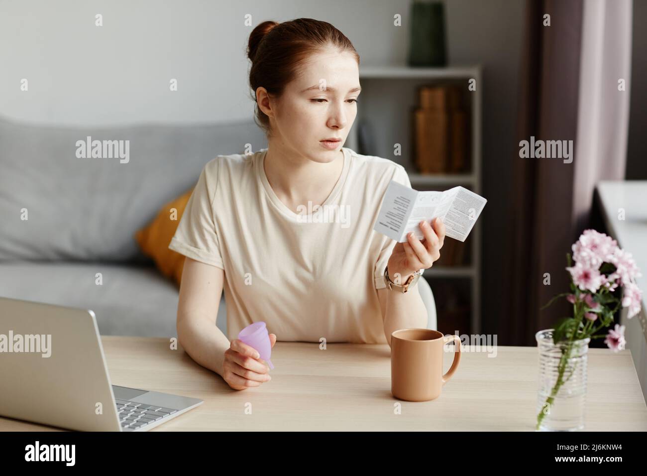 Ritratto di giovane donna caucasica lettura manuale di istruzioni per la tazza mestruale a casa in minimo interno, spazio copia Foto Stock