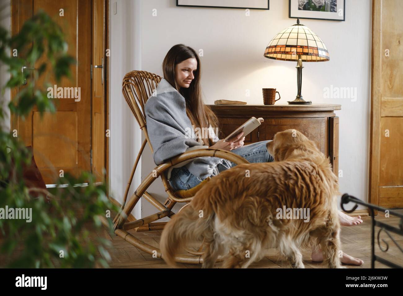 Giovane donna che legge libro con cane Golden Retriever, seduta su sedia a dondolo. Atmosfera accogliente casa, vita lenta, persone con animali domestici. Casa d'epoca Foto Stock