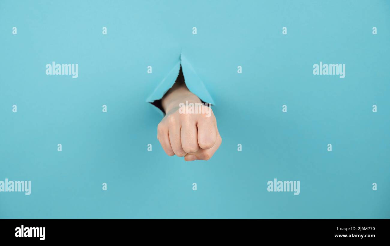 La mano di una donna che sporge da un buco da uno sfondo blu mostra un pugno. Foto Stock