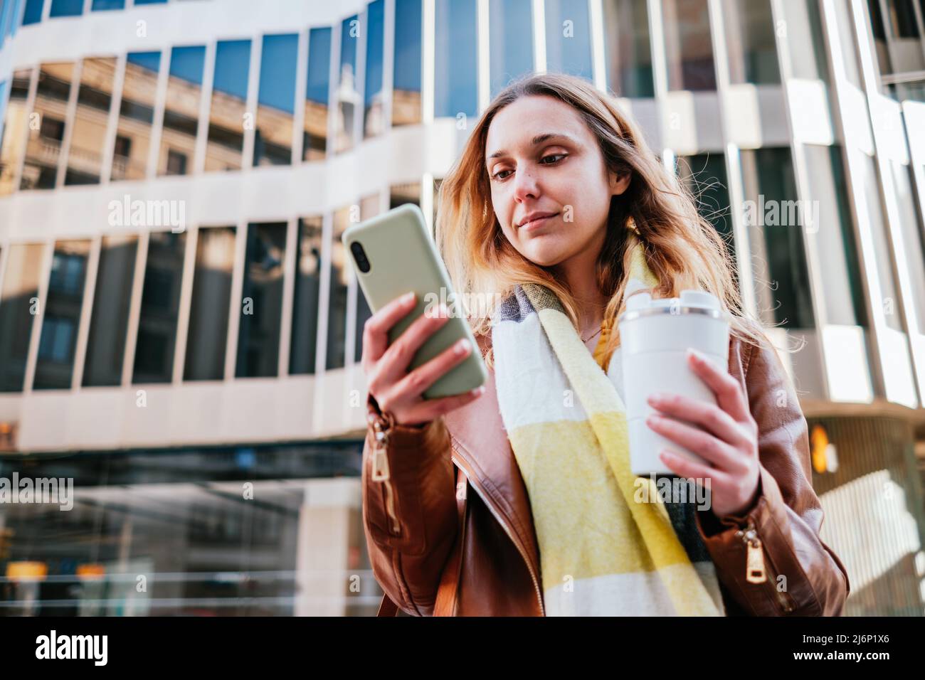 Giovane femmina con thermo tazza di caldo takeaway caffè messaggi di testo sul cellulare mentre si trova vicino a un edificio moderno sulla strada Foto Stock