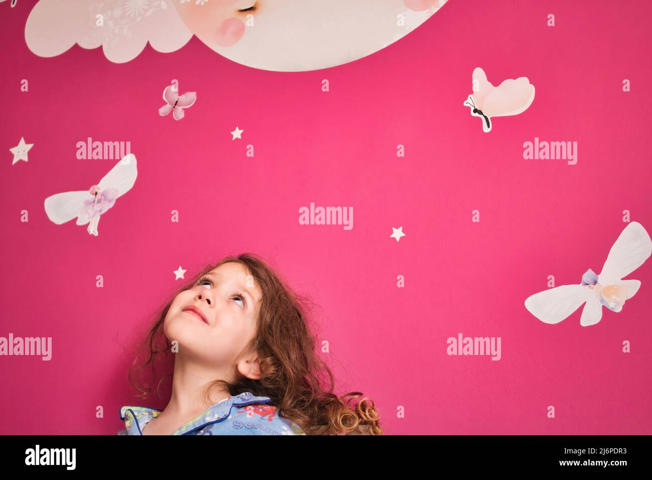 Giovane ragazza carina che indossa pigiami guardando verso l'alto un muro rosa con le stelle della decalcomania, la luna e le fate Foto Stock