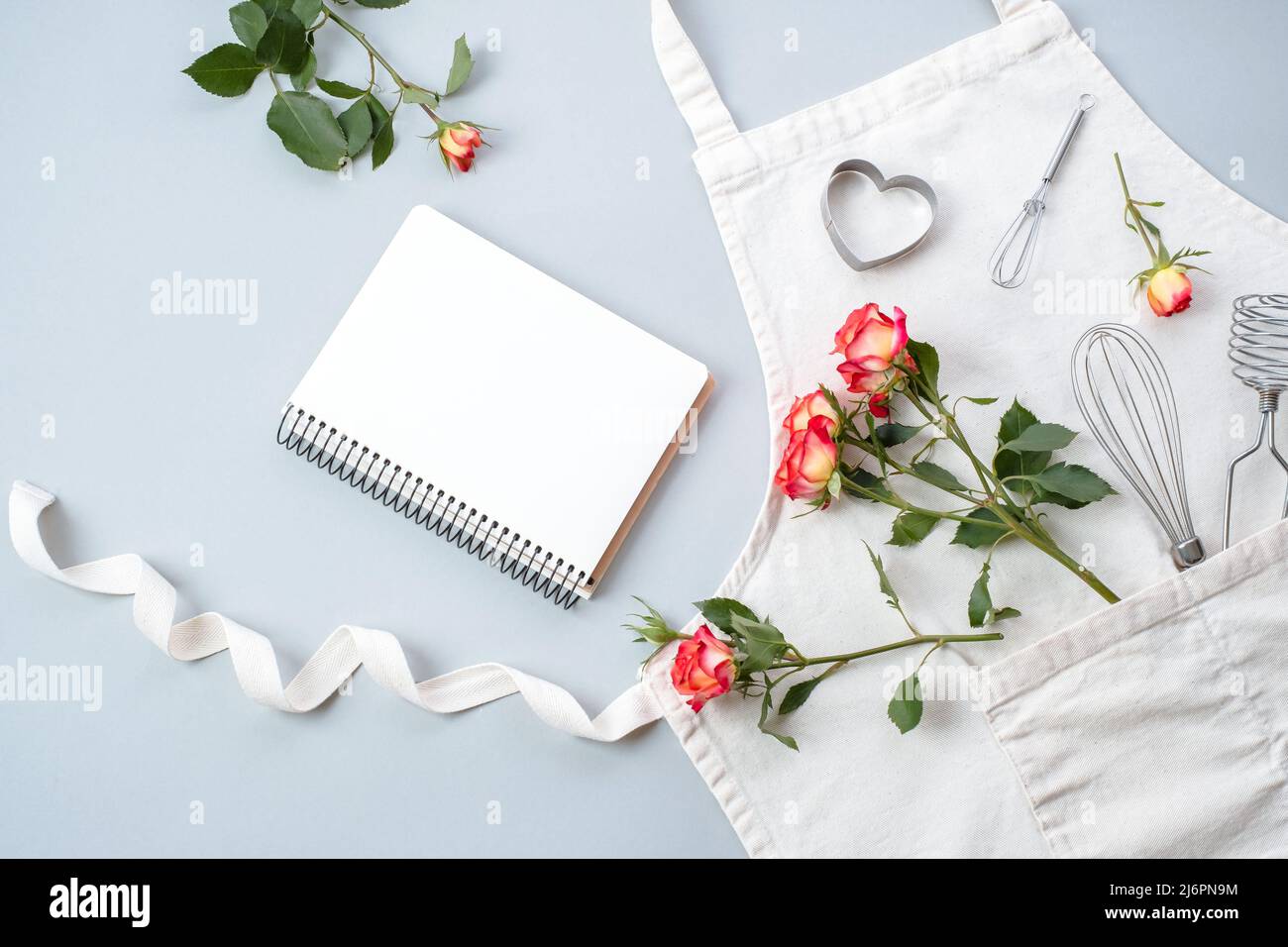 Libro da cucina in bianco, accessori da forno con grembiule con fiori rose su sfondo grigio, piatto, vista dall'alto. Ricette festive di cottura concetto. Foto Stock