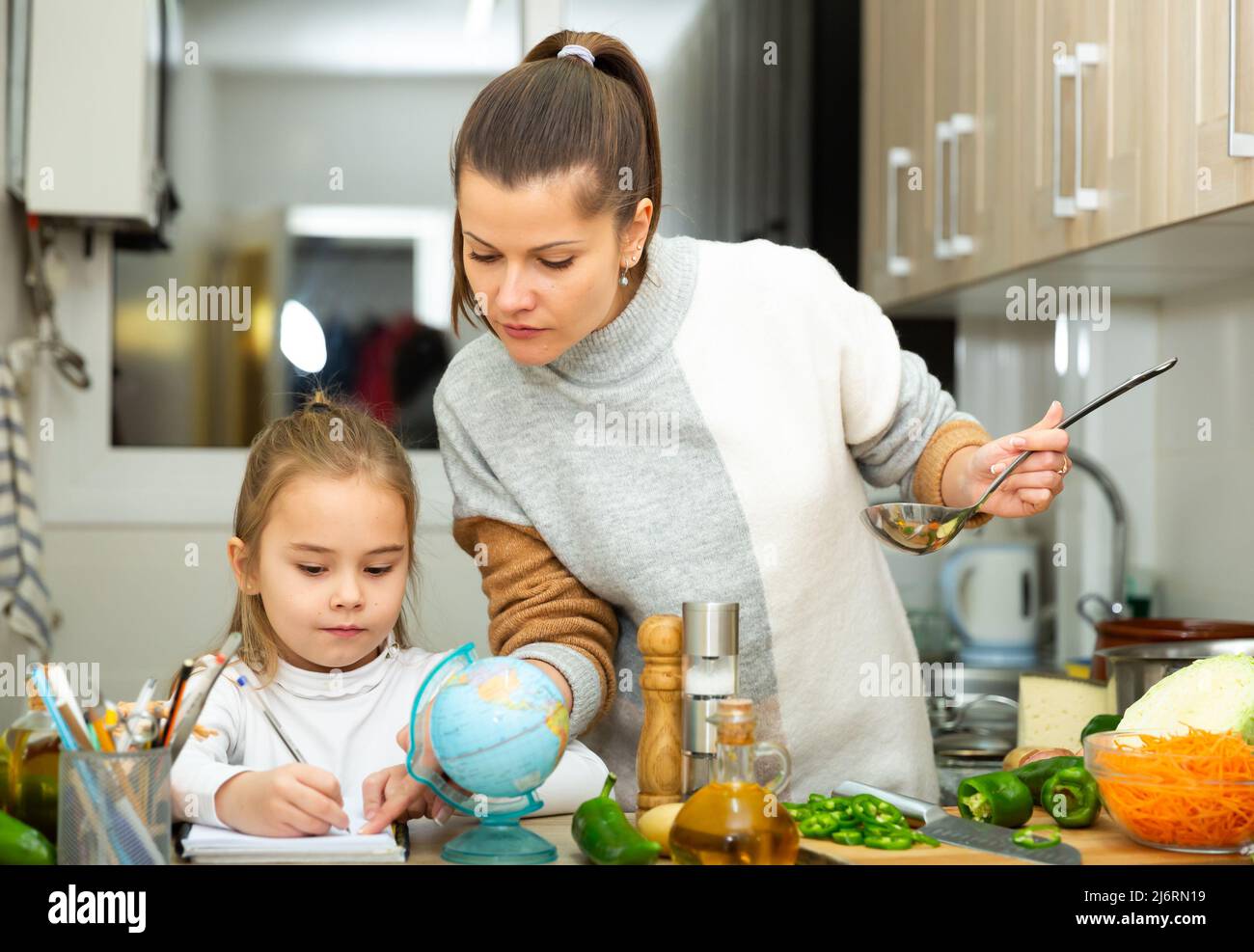 La madre che cucinava e la figlia piccola che faceva i compiti a scuola Foto Stock
