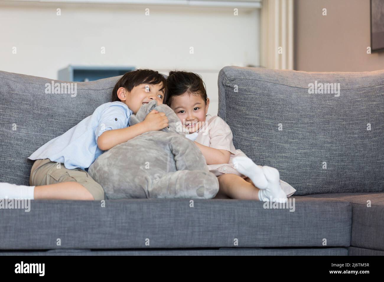 Una bambina e un ragazzino giocano felicemente con una bambola di elefante ripiena sul divano del soggiorno - foto di scorta Foto Stock