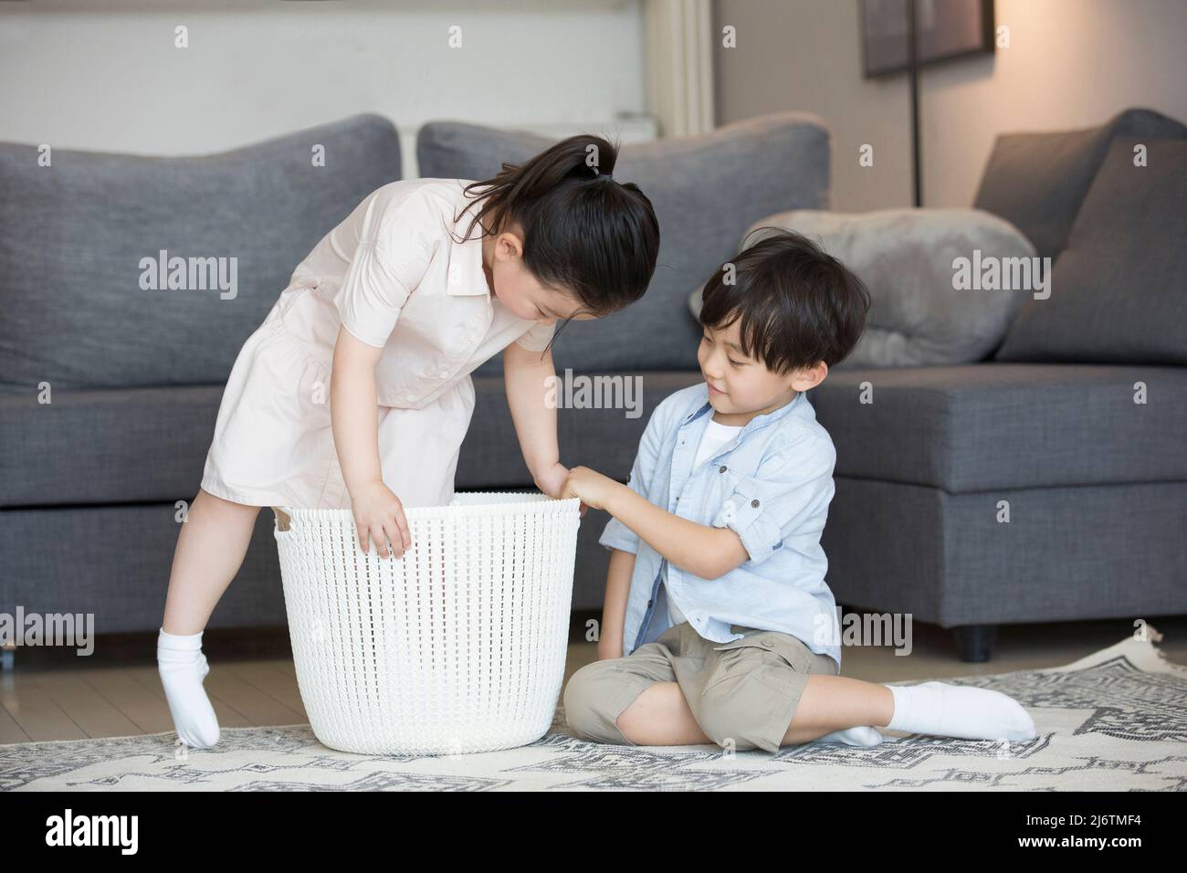 Dal divano del soggiorno, una bambina è pronta a sedersi nel cesto di vestiti e il ragazzino la sta aiutando - foto di scorta Foto Stock