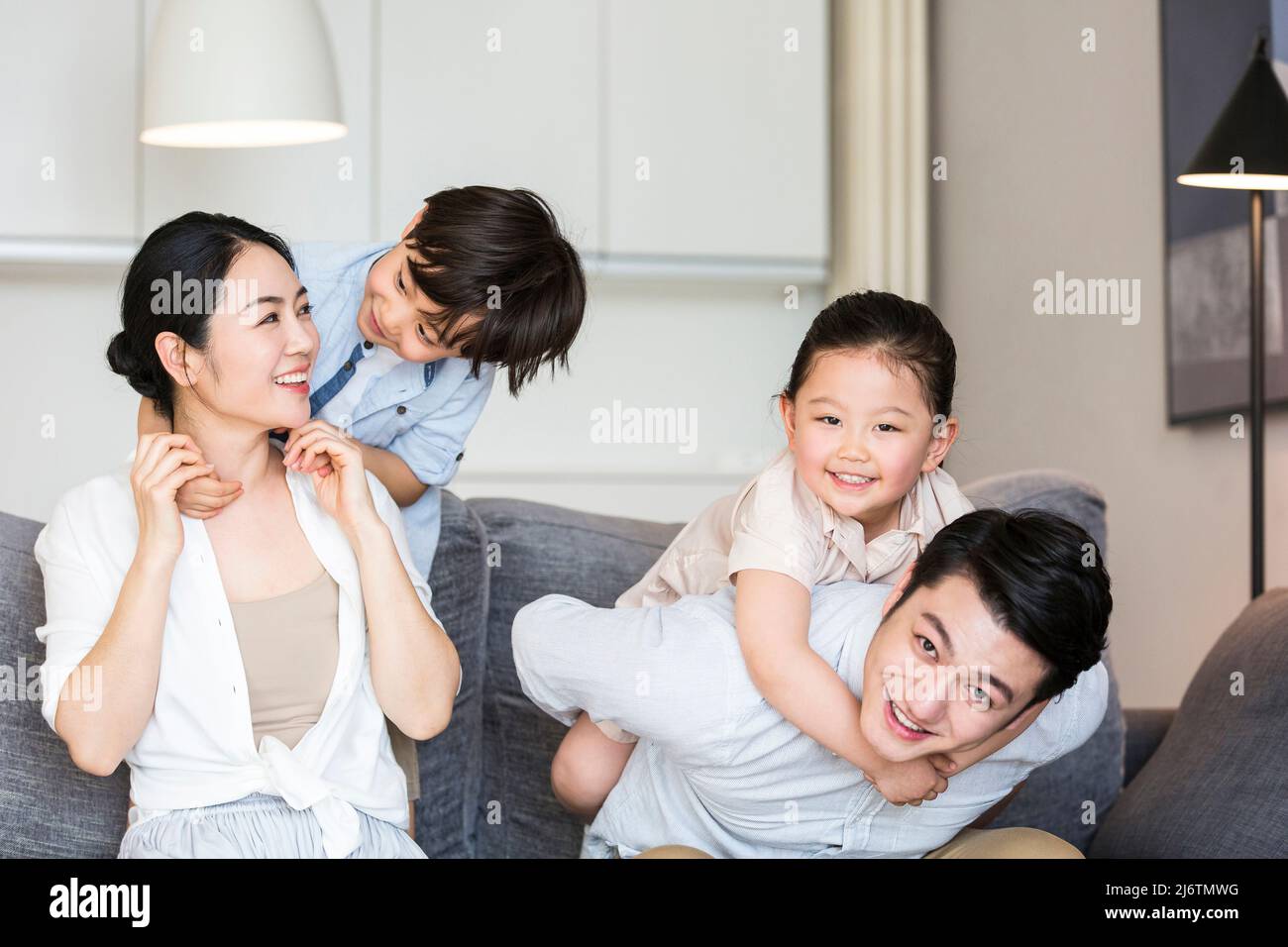 Il padre porta la figlia sulla schiena, il figlio abbraccia la madre da dietro, e la famiglia armoniosa gioca sul divano - foto di scorta Foto Stock