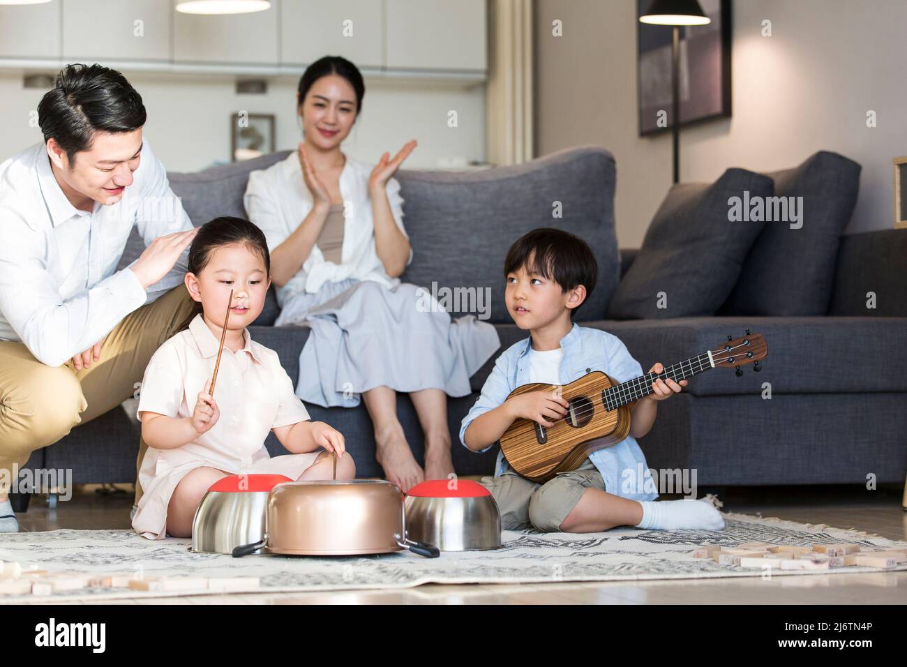 Un ragazzo e una ragazza si siedono sul tappeto giocando mischievously su ukulele e utensili da cucina, mentre i genitori guardano in ammirazione felice - foto di scorta Foto Stock