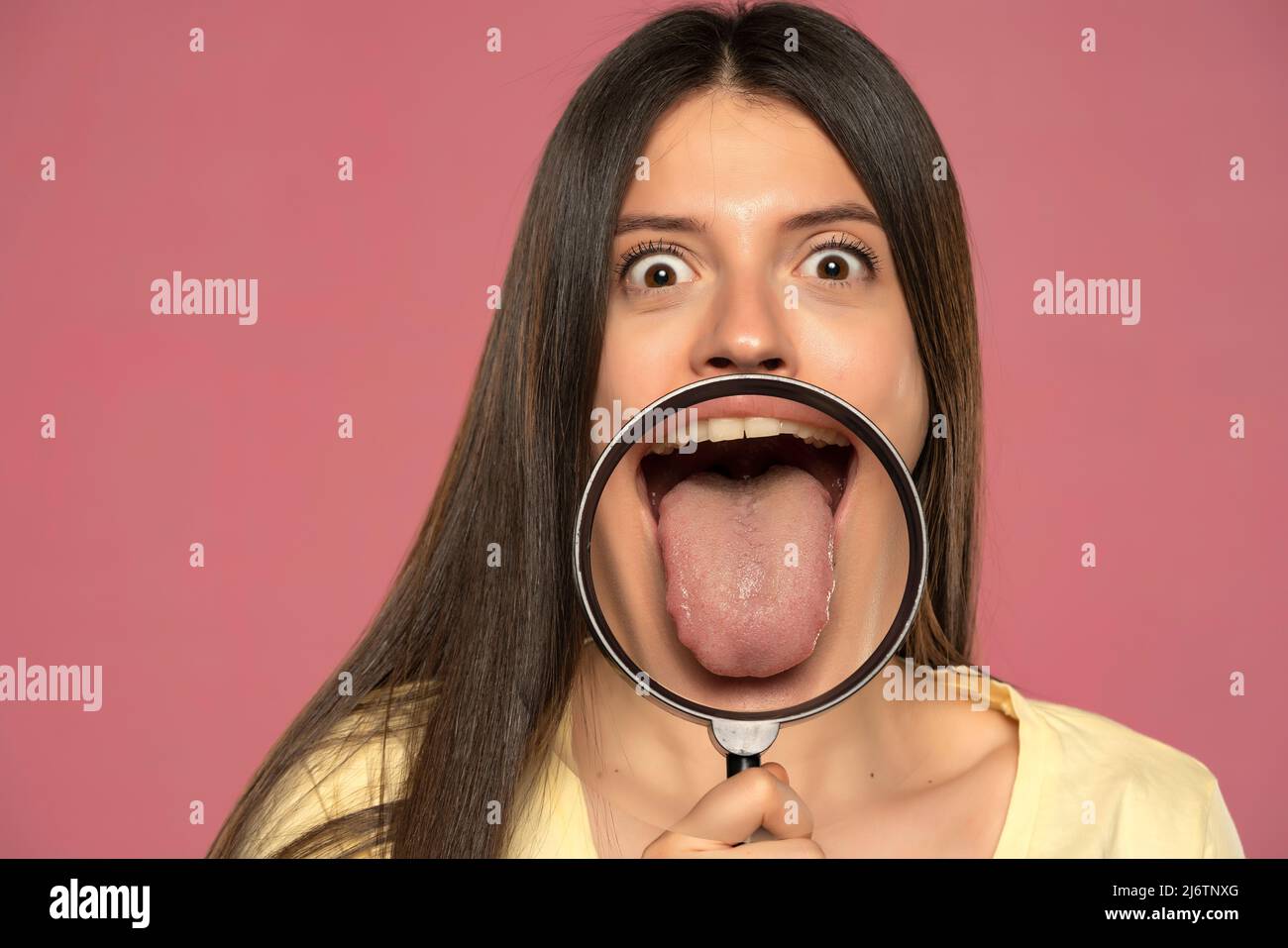 Giovane donna felice che mostra la sua lingua sana attraverso una lente di ingrandimento su uno sfondo rosa Foto Stock