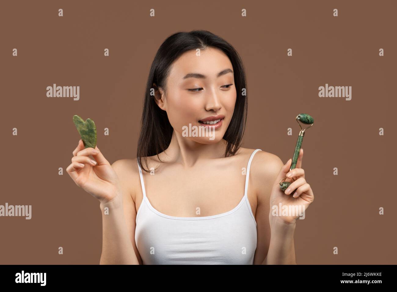 Giovane donna coreana che tiene bellezza viso gua sha e jade rullo di quarzo, scegliendo cosa usare, sfondo marrone Foto Stock