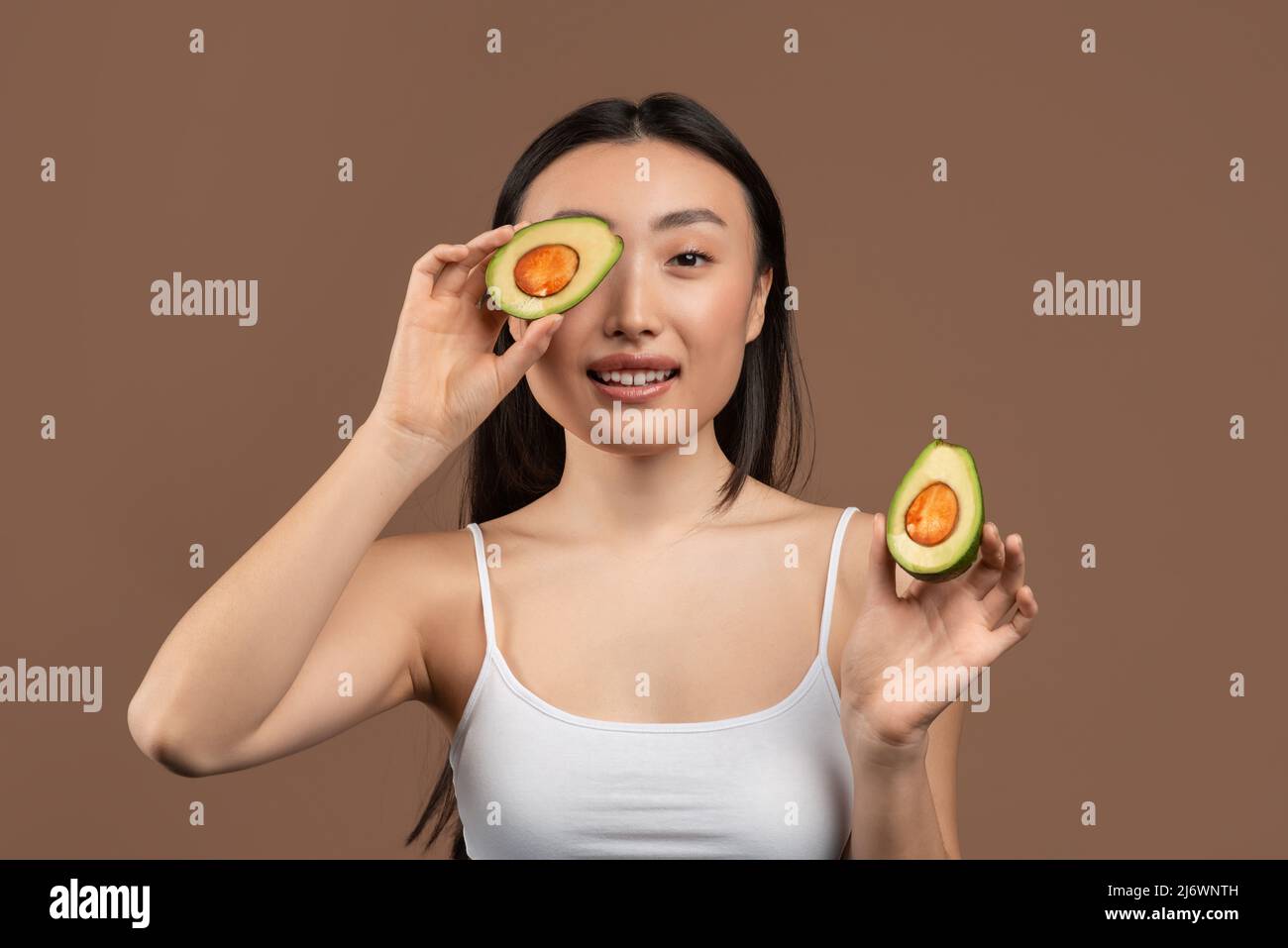 Bellezza e anti-invecchiamento ricette biologiche concetto. Bella donna coreana che copre l'occhio con metà di avocado, sfondo marrone. Foto Stock