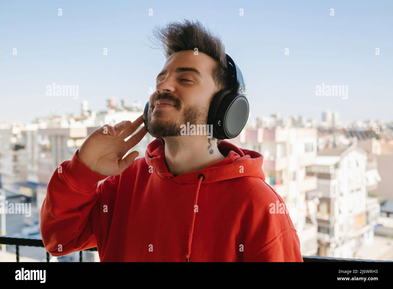 Buon giovane uomo con cappuccio rosso che ascolta la musica con le cuffie. Ascoltare la sua musica preferita. Cuffie che ascoltano e sorridono mentre si è in piedi Foto Stock