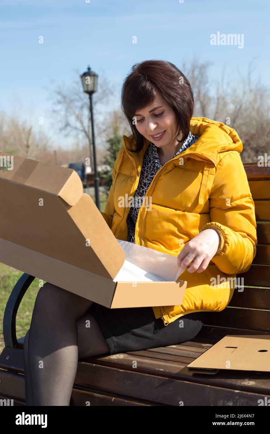 Una donna felice bruna in una panca giallo brillante giacca con un sorriso apre una scatola con un nuovo portatile. Foto Stock