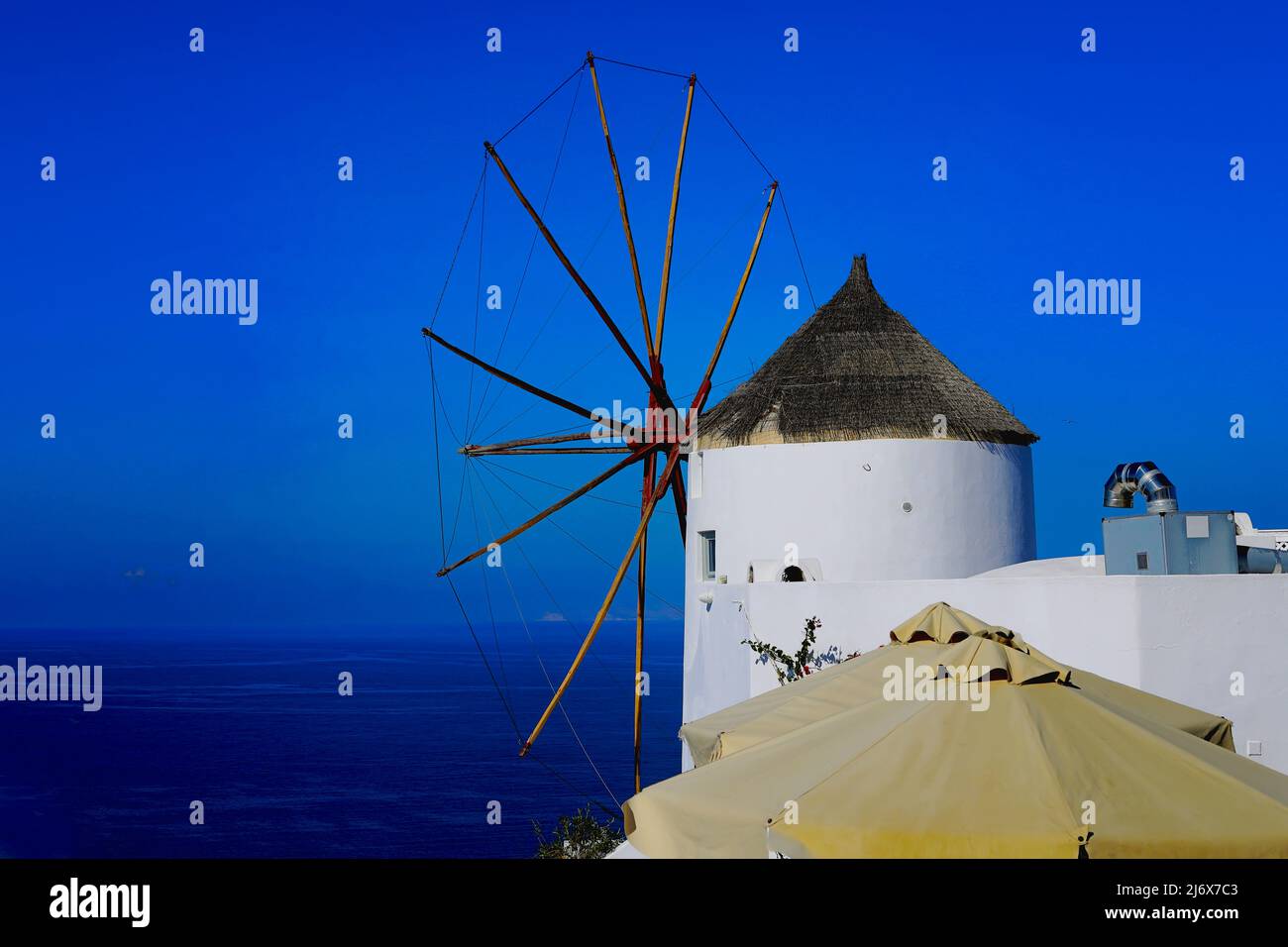 Oia, Santorini, Isole Greche, Grecia - Mulino a vento Pretty imbiancato con ombrelloni in primo piano arroccato sulla scogliera in un sole abbagliante e luminoso Foto Stock
