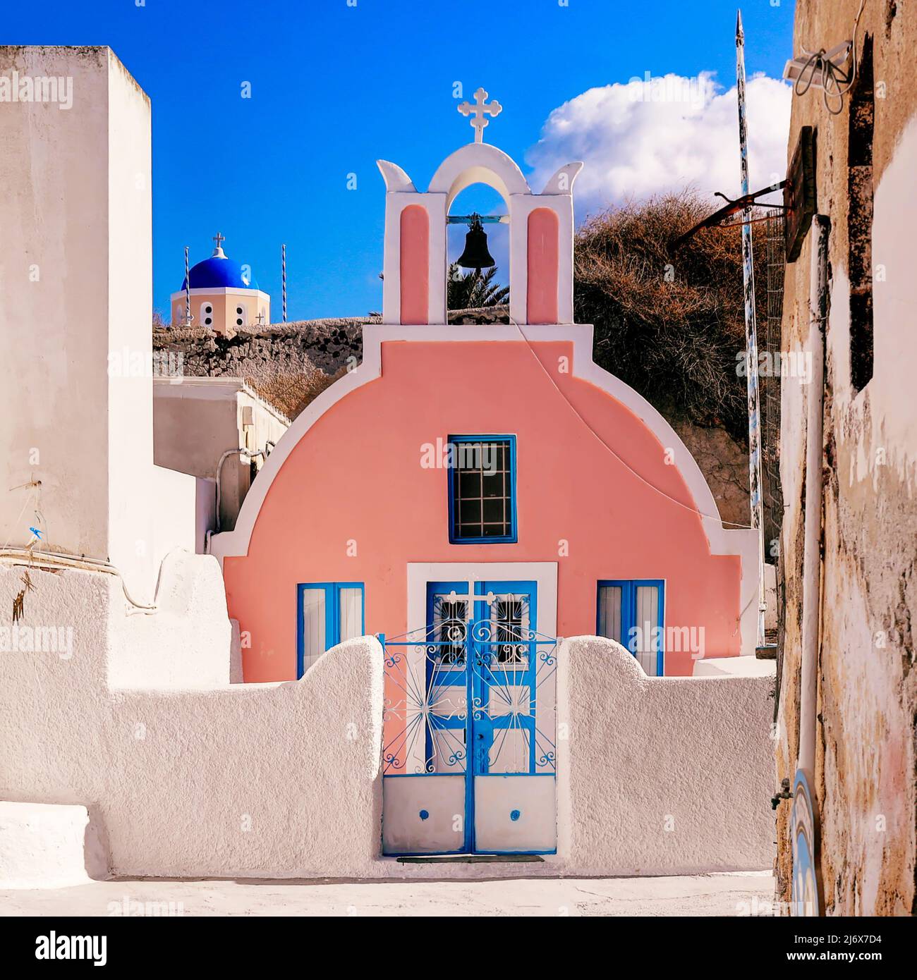 Oia, Santorini, Isole Greche, Grecia - Chiesa greco-ortodossa di colore rosa Pretty con campanile e chiesa a cupola blu alle spalle in una luce del sole abbagliante Foto Stock