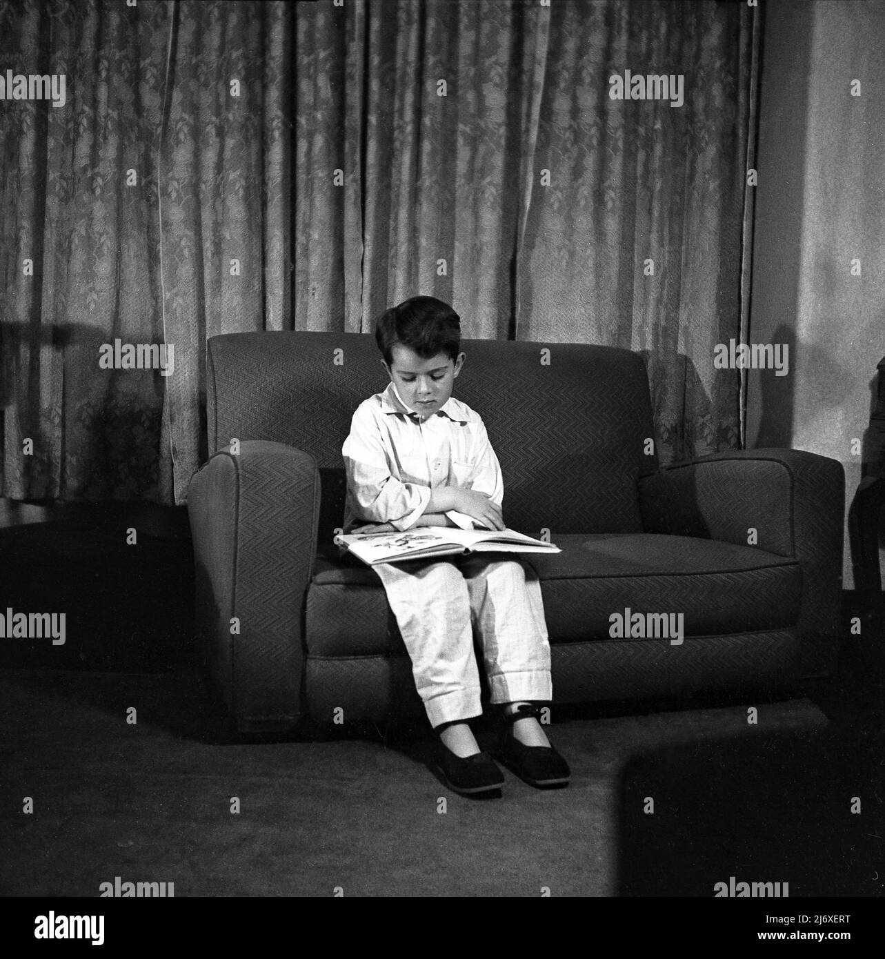 1950s, storico, giovane ragazzo nei suoi pigiami e pantofole seduti su un piccolo divano guardando un libro di foto prima di andare a dormire, Inghilterra, Regno Unito. Foto Stock
