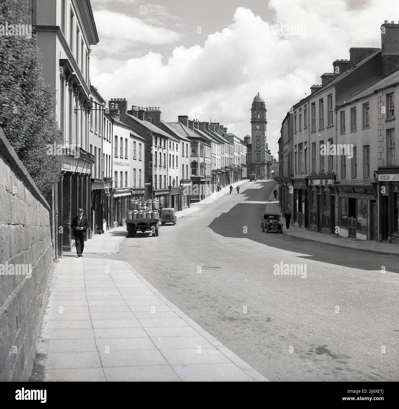 1950s, Historical, Enniskillen, Irlanda del Nord, Regno Unito, mostra una strada alta vicina deserta di con solo un paio di locali che camminano lungo il marciapiede. Un camion posteriore aperto con le urne del latte è parcheggiato su. Risalente al 1901, la torre dell'orologio del municipio è visibile all'estremità della strada. I nomi nei negozi includono un bar, William Blake, un macellaio e un portavetrini Peter Burns. La targa su una delle vetture parcheggiate nella strada deserta, il 2202, mentre sull'altro lato della strada, un'altra auto, di un'epoca ancora precedente è parcheggiata e questo ha il numero HMT 275. Foto Stock