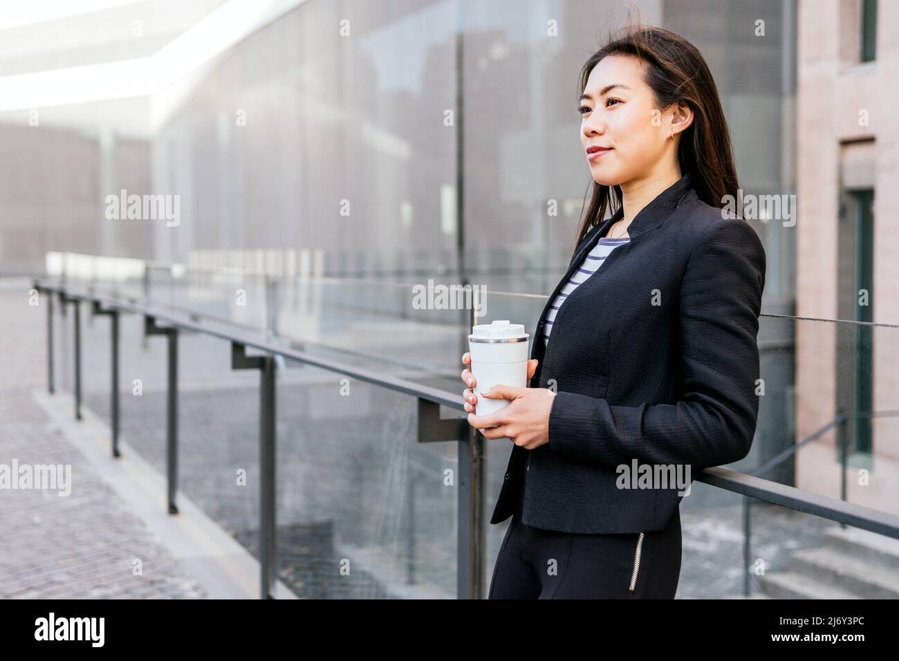 Giovane e ottimista impiegato asiatico in ufficio con tuta formale in piedi con bicchiere in acciaio riutilizzabile per bevande da asporto appoggiate sulla ringhiera nel centro della città Foto Stock