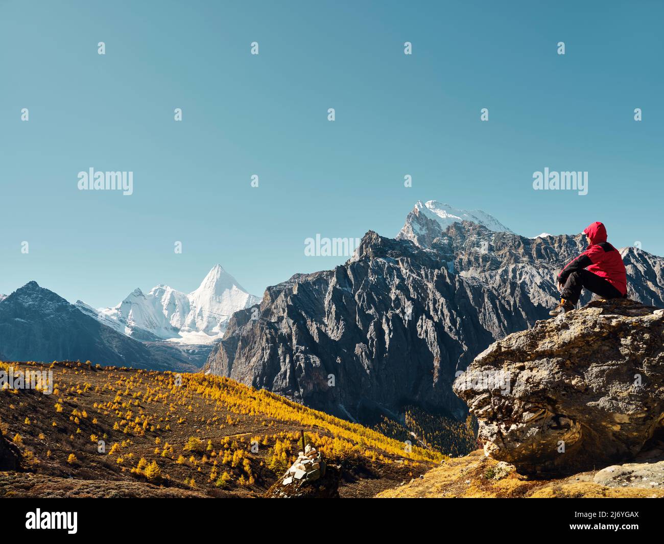 Uomo asiatico seduto in cima alla roccia guardando Yangmaiyong (o Jampayang in tibetano) picco di montagna in lontananza in Yading, Daocheng County, Sichuan Pr Foto Stock