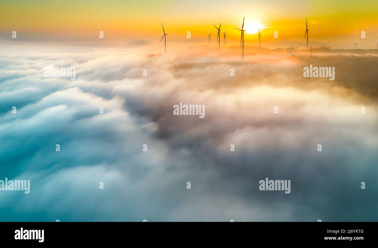 Vista aerea collina in cima all'alba con nebbia che copre un piccolo villaggio nella valle, bei poli di energia eolica che si innalzano in alto per accogliere una tranquilla nuova giornata Foto Stock