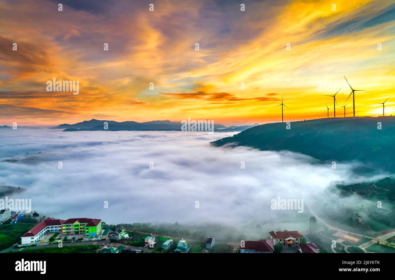 Vista aerea collina in cima all'alba con nebbia che copre un piccolo villaggio nella valle, bei poli di energia eolica che si innalzano in alto per accogliere una tranquilla nuova giornata Foto Stock