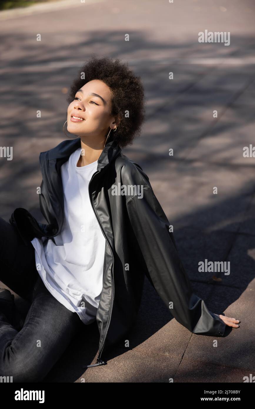 compiacente donna afroamericana in orecchini a cerchio e giacca seduta su asfalto Foto Stock