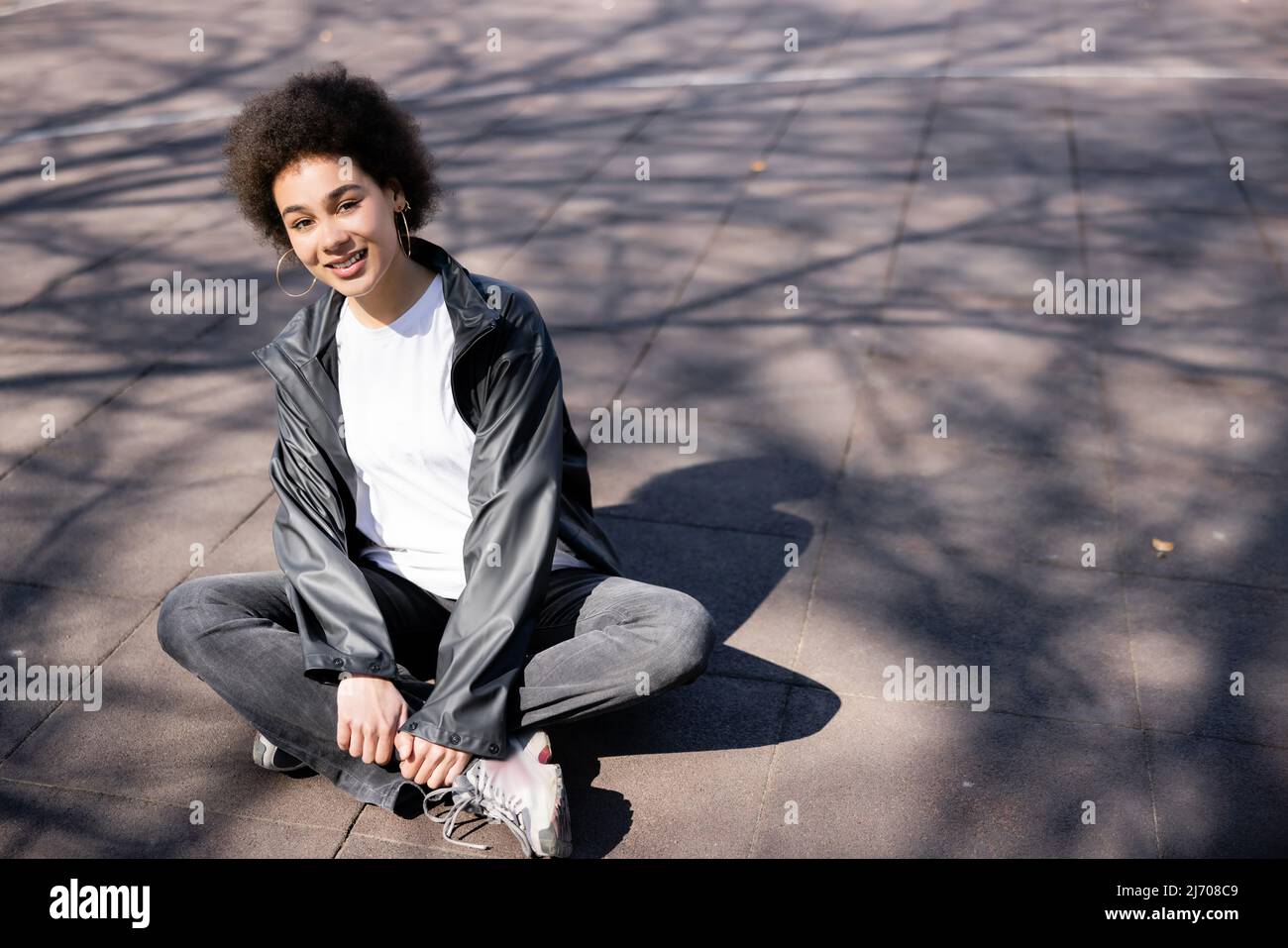 intera lunghezza di felice afroamericana donna in giacca seduta con gambe incrociate su asfalto con ombre Foto Stock