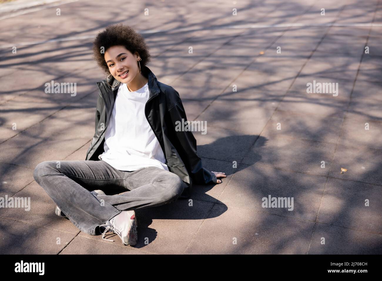 intera lunghezza di sorridente donna afroamericana in giacca seduta con gambe incrociate su asfalto con ombre Foto Stock