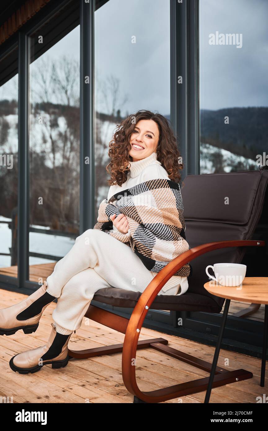Giovane donna curly che riposa sulla terrazza della moderna casa di fienile in montagna. Felice turista femminile in plaid seduta su sedia, godendo in nuovo cottage in inverno. Foto Stock
