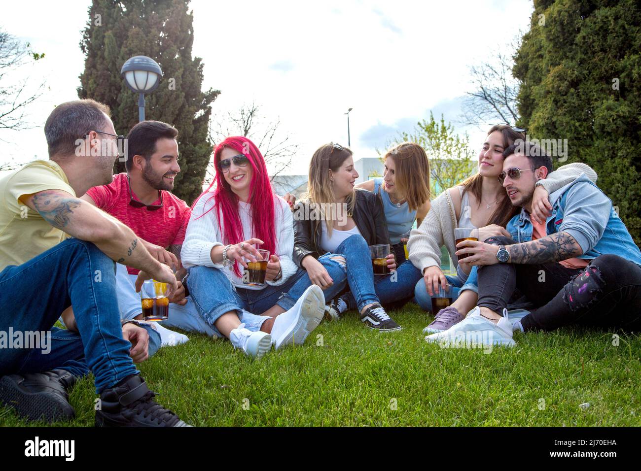 gruppo di amici seduti sull'erba che hanno un bel po' di tempo con bicchieri di bevande in mano e che parlano Foto Stock