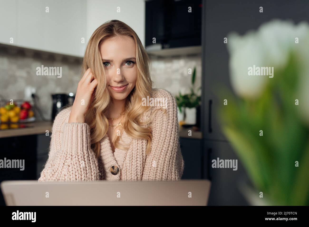 Giovane donna felice seduta in cucina al mattino e sorridente lavorando sul portatile. Ritratto di signora in abiti casual liberelancing da casa. Concetto di Foto Stock