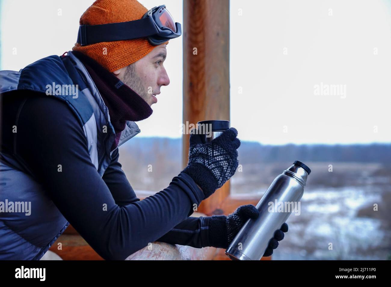 Un ragazzo in un cappello arancione in inverno beve il tè da un thermos durante una vacanza invernale in montagna. Si trova sul balcone di una casa di legno. Porta Foto Stock
